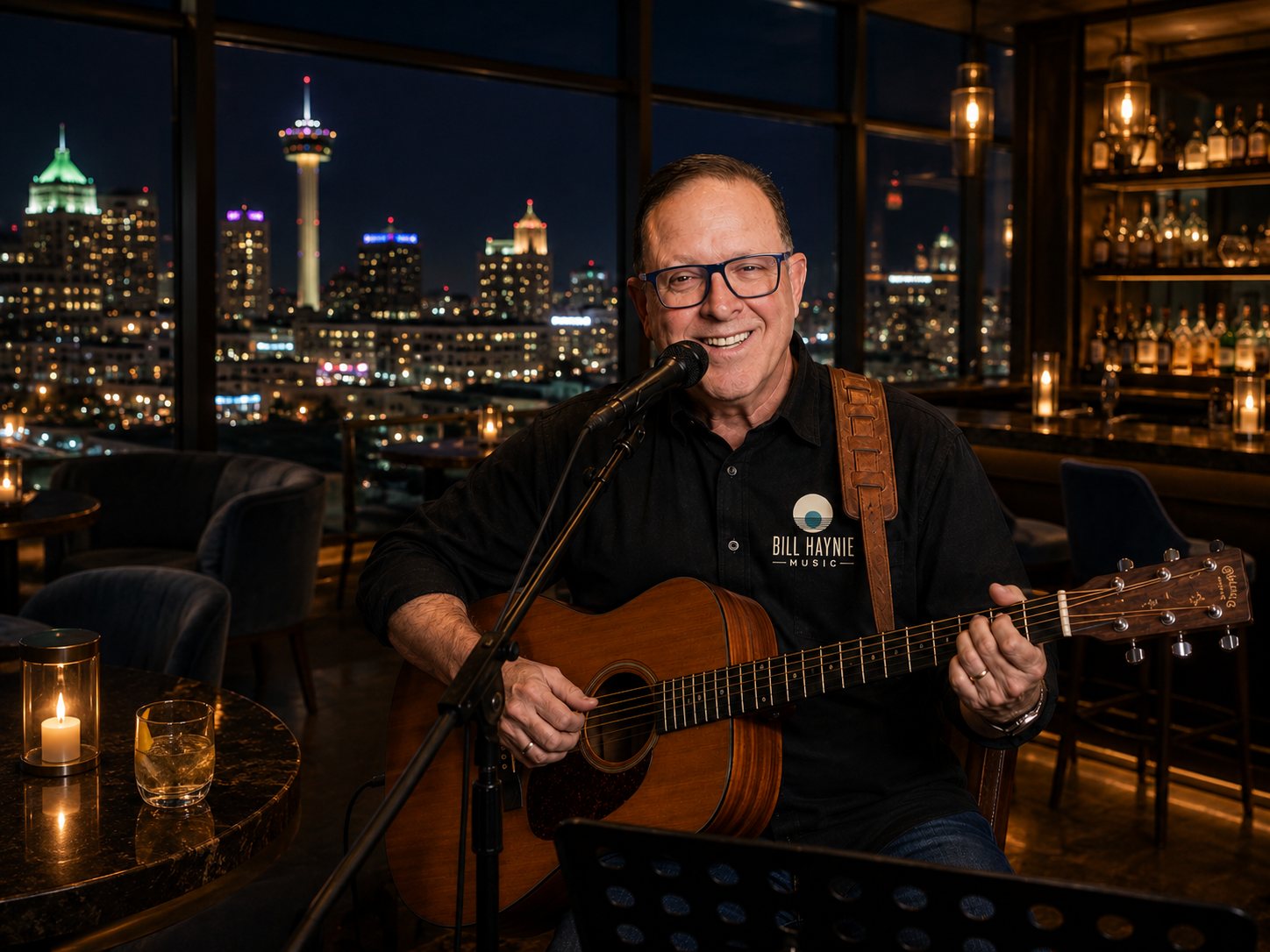 Bill Haynie with his acoustic guitar at a rooftop venue overlooking the San Antonio skyline at night